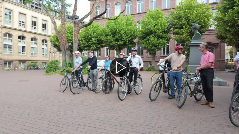 Gruppe von Menschen mit Fahrrädern auf einem gepflasterten Platz vor einem Gebäude.