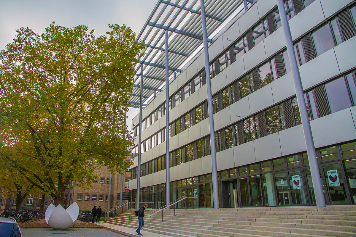 Modernes Institutsgebäude mit Glasfassade, einer großen Treppe und einem herbstlichen Baum davor.